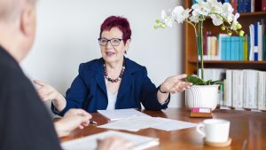 Die Fachanwältin für Arbeitsrecht berät Mandanten. Die Anwältin sitzt am Besprechungstisch. Darauf liegen Unterlagen. Sie berät einen Mandanten, der nur von hinten zu sehnen ist.
